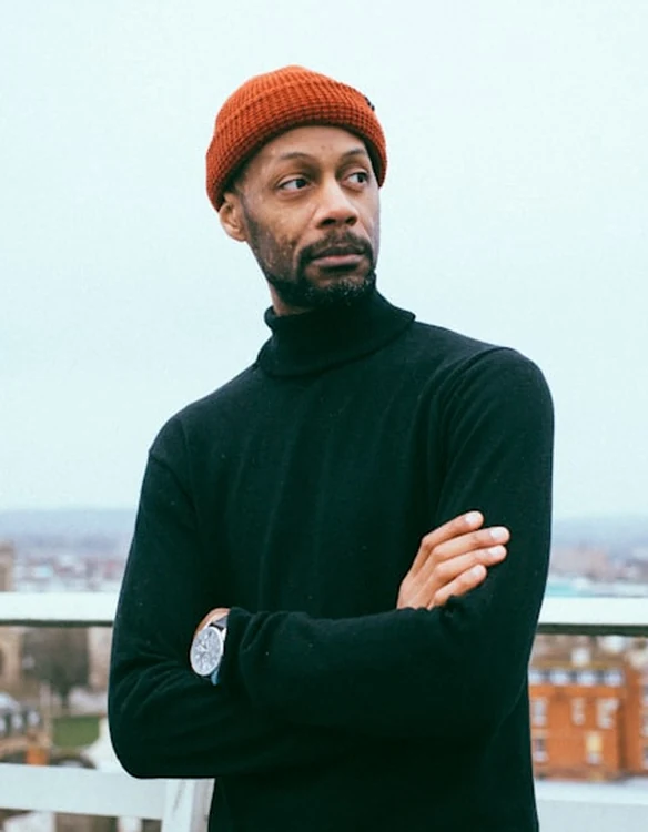 Portrait of a man in an orange beanie and black turtleneck on a city rooftop.