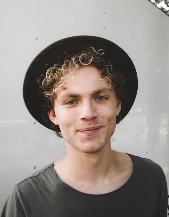 Portrait of a curly-haired man in a wide-brimmed hat against a light grey wall.