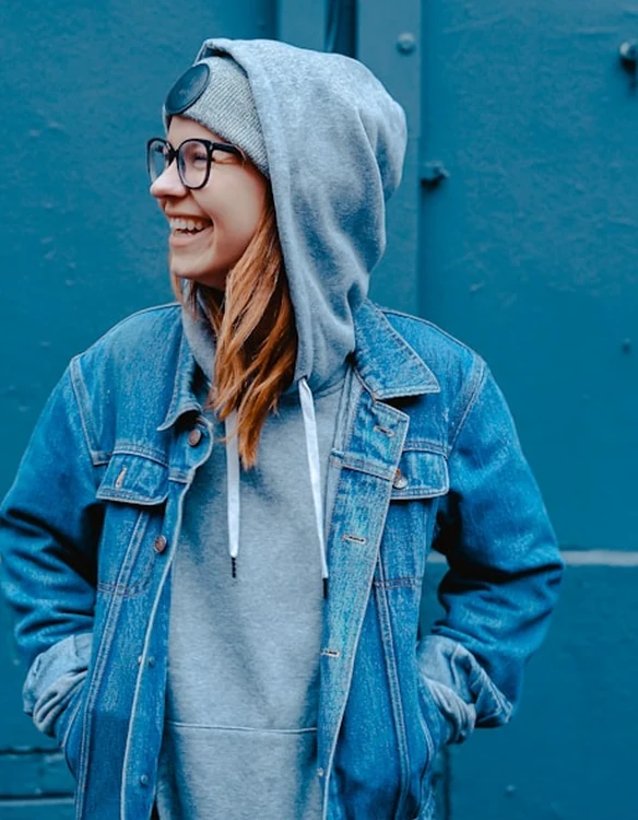 Portrait of a woman in a denim jacket and grey hoodie laughing beside a blue wall.