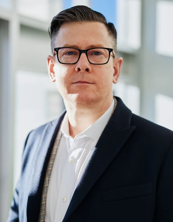 Portrait of a man in dark-rimmed glasses and a navy blazer in a bright office.