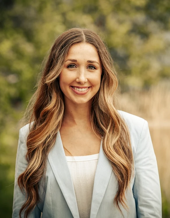 Portrait of a woman in a pale blue blazer smiling outdoors in front of trees.