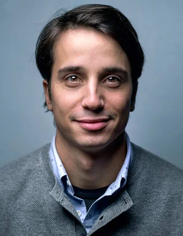Studio portrait of a man in a grey sweater over a blue collared shirt.